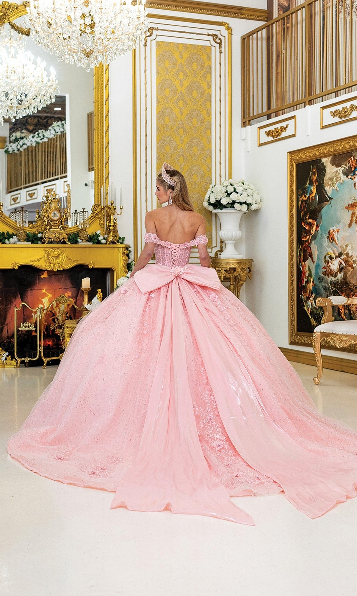 A woman wears the Quinceanera Dress 5056 By Dancing Queen, a large pink gown with a sheer corset bodice and 3D flowers, standing with her back to the camera in an ornate gold-accented room with a chandelier and classical art.
